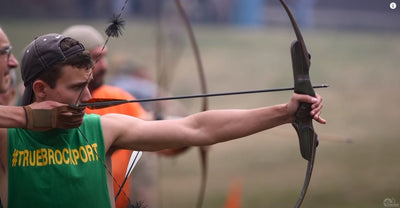 A man holding his traditional recurve bow outside at full draw and anchoring below his cheek bone.