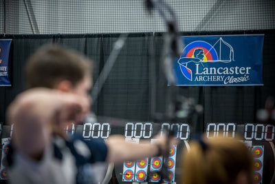 An archer is at full draw, while the focus is on the Lancaster Archery Classic sign in front of him.