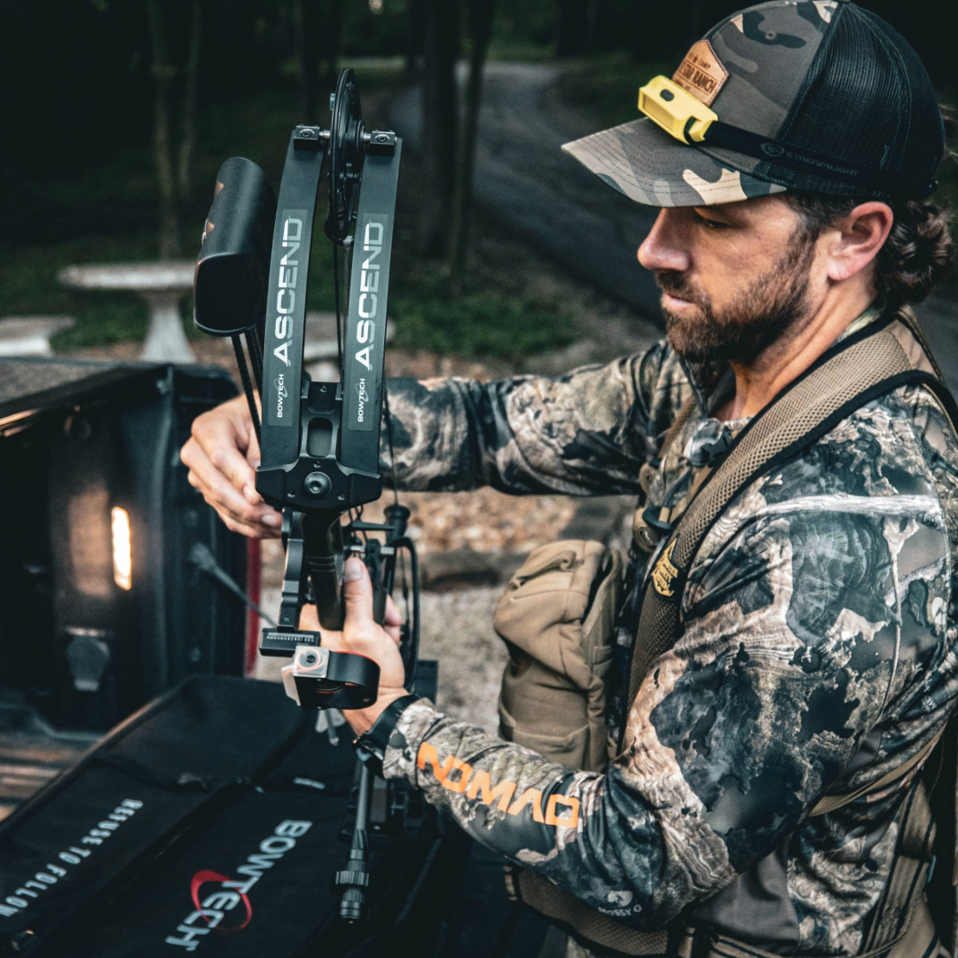 A man in camouflage gear is adjusting a bow on a truck bed outdoors.
