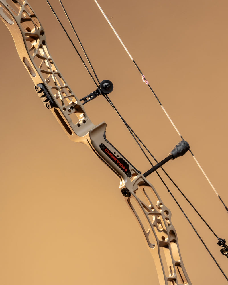 Close-up of a compound bow with a tan and black riser, black cables, and a black stabilizer against a solid tan background.