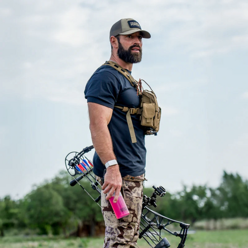 A man wearing a binocular harness holding a compound bow in an outdoor setting.