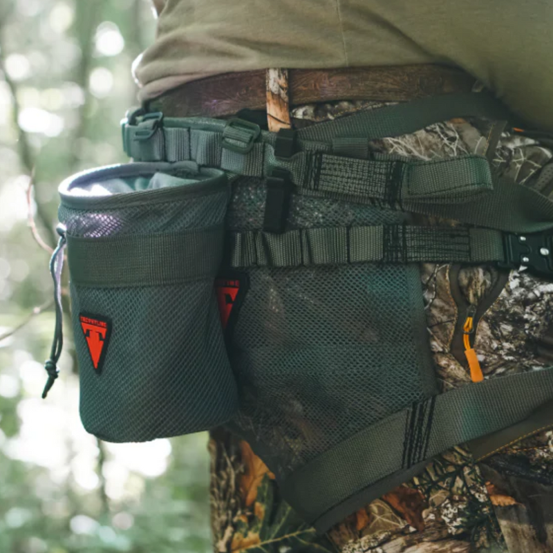 A close-up of a camouflage hunting waist harness with a dark green mesh side pouch, worn over camouflage pants in a forest setting.